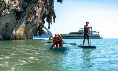 Ajao: Duas pessoas em um caiaque e uma em uma prancha de stand up paddle perto de um penhasco, o iate ao fundo