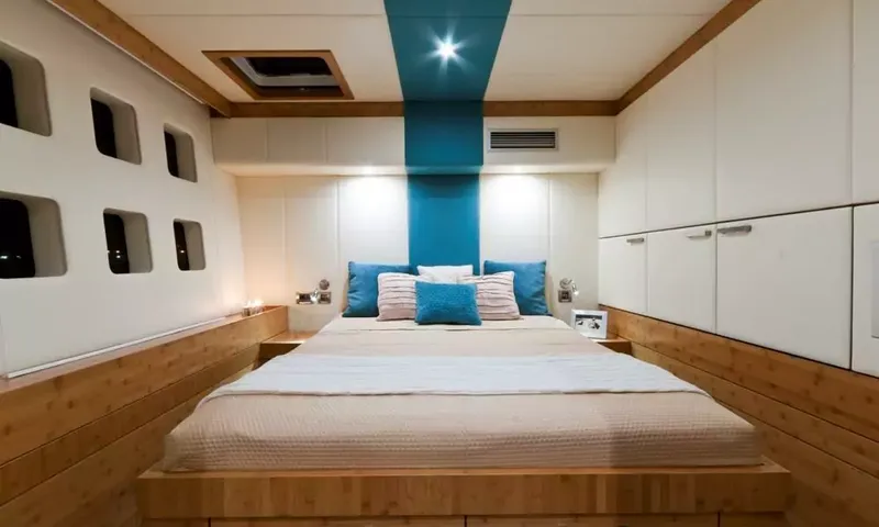 Anini: Double bed on a wooden base with blue cushions, hull windows and white cupboards
