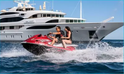 Galaxy: Duas pessoas em um jet ski vermelho levantando respingos ao lado do iate ancorado