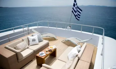 Ithaki: Lounge del ponte superiore con divani in vimini, tavolino e lettini prendisole affacciati su un mare calmo, bandiera al vento