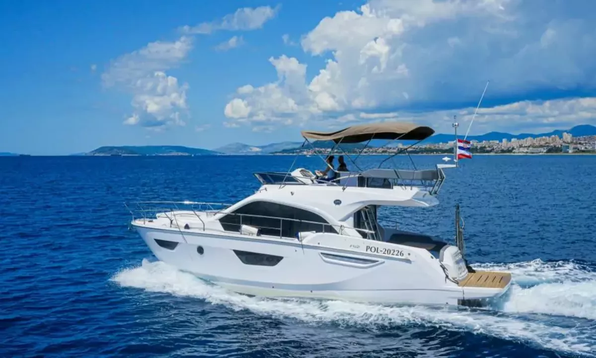 Jupika II: Yacht à moteur blanc avec flybridge naviguant sur une mer bleu profond, avec une ville côtière et des montagnes en arrière-plan
