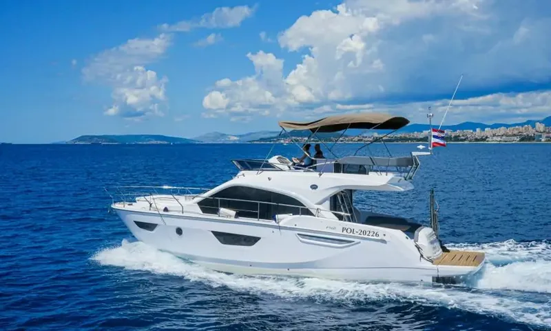 Jupika II: Yacht à moteur blanc avec flybridge naviguant sur une mer bleu profond, avec une ville côtière et des montagnes en arrière-plan