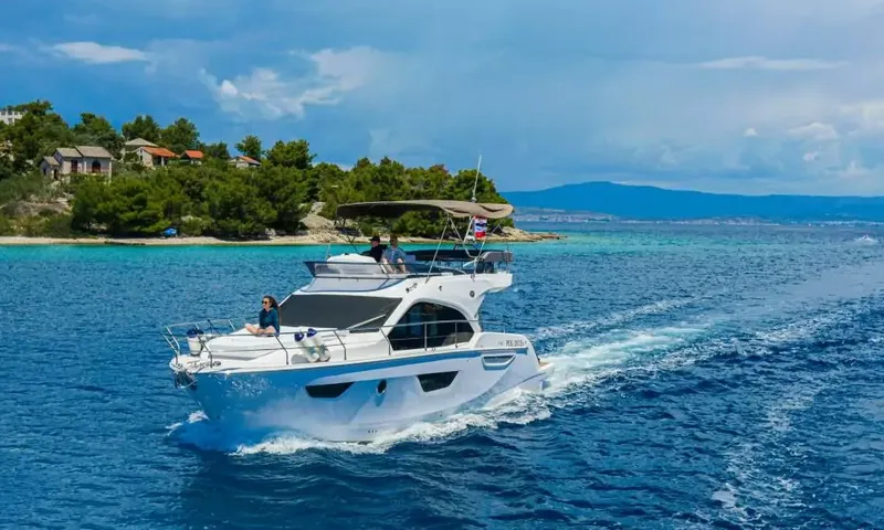 Jupika II: Yacht à moteur en route près du rivage boisé d'une île, avec des invités à l'avant et sur le flybridge