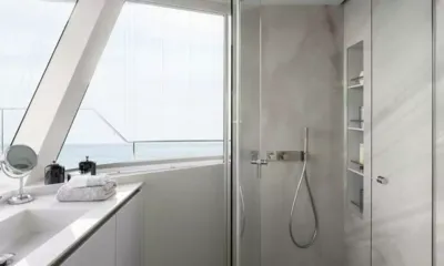 Mayrilou: Banheiro iluminado com chuveiro de vidro, bancada branca, toalhas dobradas e janela inclinada com vista para o mar