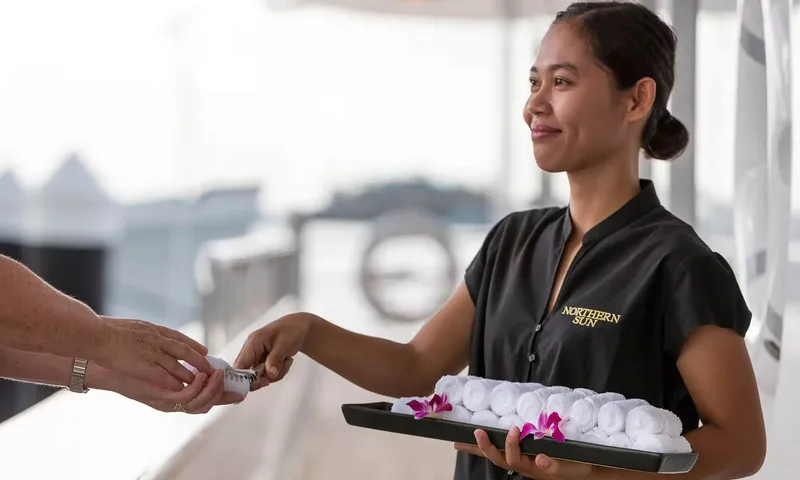 Northern Sun: Crew member in black uniform offering rolled towels from a tray decorated with orchids