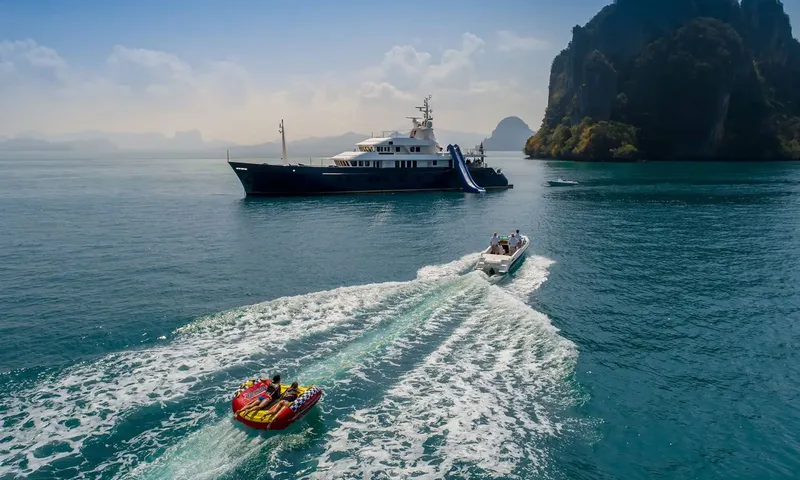 Northern Sun: Yacht with a deck slide at anchor while a small boat tows an inflatable tube near cliffs