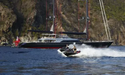 Seahawk: Zwei Personen auf Jetskis fahren an einer vor Anker liegenden Segelyacht mit dunklem Rumpf nahe einem grünen Hügel vorbei