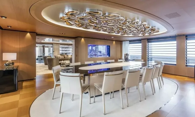 Suerte: Oval dining table with white chairs under a silver ceiling sculpture, aquarium in the background