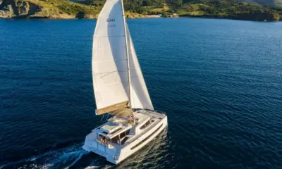 Happy Fifty: Catamarano a vela a vele spiegate su acqua blu intenso vicino a una costa verde e collinosa