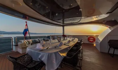 Aiaxaia: Set oval dining table on aft deck with flag and sunset over the water