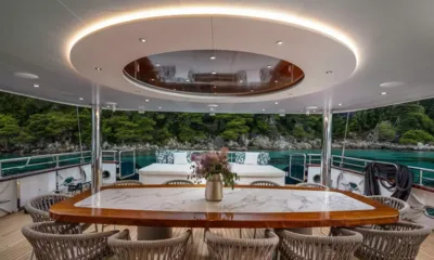 Nocturno: Mesa de jantar externa coberta com tampo de mármore e cadeiras trançadas, com mar e margem arborizada ao fundo