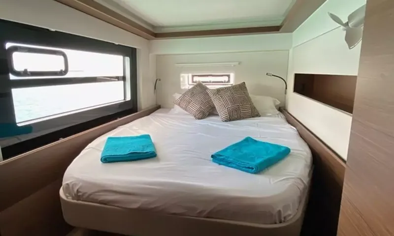 Sea Spirit 3: Cama de casal com almofadas estampadas e toalhas azuis ao lado de uma janela no casco