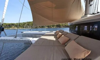 Breiz-Ile: Foredeck sunpads with cushions under a shade sail beside trampoline nets, wooded coast behind