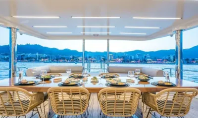 Deep Water: Mesa de jantar no convés de popa coberto com cadeiras de vime e uma cidade iluminada à beira-mar ao entardecer