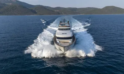 Go: Yacht se dirigeant vers la caméra à vive allure avec des embruns à l'étrave, collines vertes à l'arrière-plan
