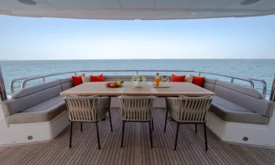 Legende: Mesa de jantar de teca no convés de popa com cadeiras trançadas e sofá embutido voltados para o mar aberto