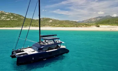 One Planet: Catamarã azul-escuro ancorado em águas turquesa em frente a uma praia de areia com colinas verdes
