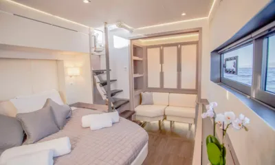 D2: Cabine iluminada com cama de casal, toalhas, um sofá pequeno, escadas e janela com vista para o mar