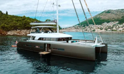 La Gatta: Catamarã a vela ancorado perto de uma colina verde e de uma cidade costeira com a plataforma do casco abaixada