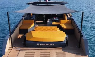 Flying Spur: Convés de popa com toldo sobre um grande colchonete de sol amarelo e sofás laterais, plataforma de teca à frente