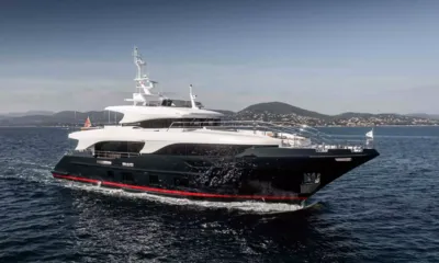 Tonic Blue: Yacht con scafo scuro e striscia rossa in navigazione davanti a colline verdi e a una cittadina costiera