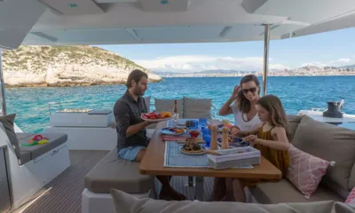 Rise: Família almoçando na mesa sombreada do cockpit, com o mar e o litoral ao fundo