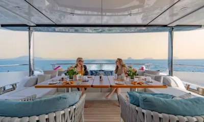 Song of Songs: Duas mulheres jantam em uma mesa posta em um convés de popa sombreado com vista para o mar