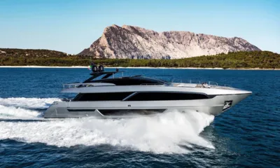 Corsaro: Yacht argentato che naviga veloce davanti a un'isola rocciosa sotto un cielo azzurro limpido