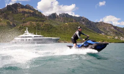 Reliance: Piloto em um jet ski azul levantando água, com um iate ancorado sob montanhas verdes