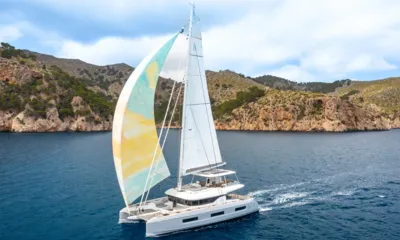 Wish: Catamarã navegando com um spinnaker pastel ao longo de uma costa rochosa e verde
