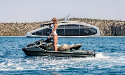 This Is It: Mulher em pé em um jet ski com o iate ancorado perto de uma margem rochosa ao fundo