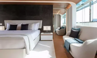 Legend: Cabina con grande letto bianco, parete scura dietro la testata e divano curvo vicino a finestre alte