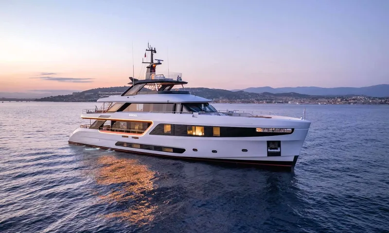 Angel: White motor yacht at anchor at dusk with lit windows and hills behind