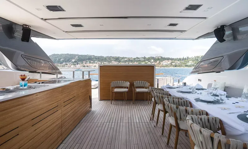 Kristo: Shaded outdoor dining table with woven chairs beside a wooden bar counter, coastline beyond