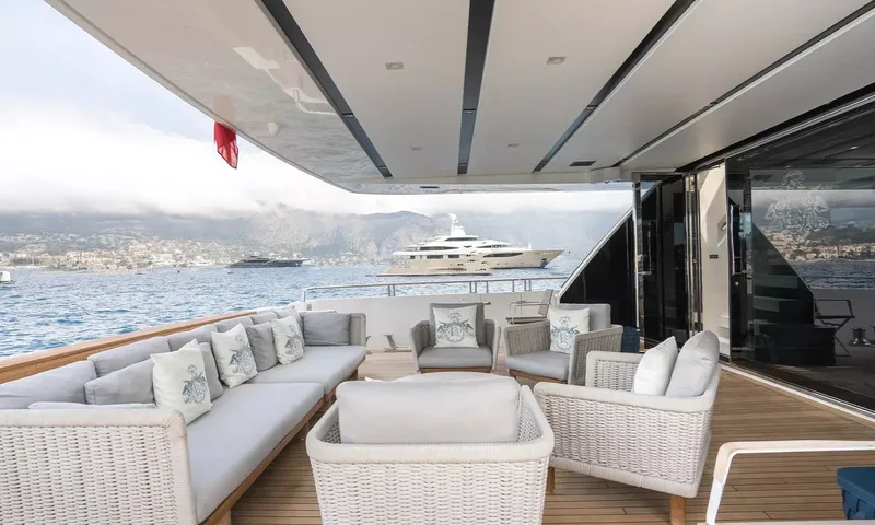 Kristo: Shaded aft deck seating with woven armchairs and sofa beside glass doors, yachts offshore