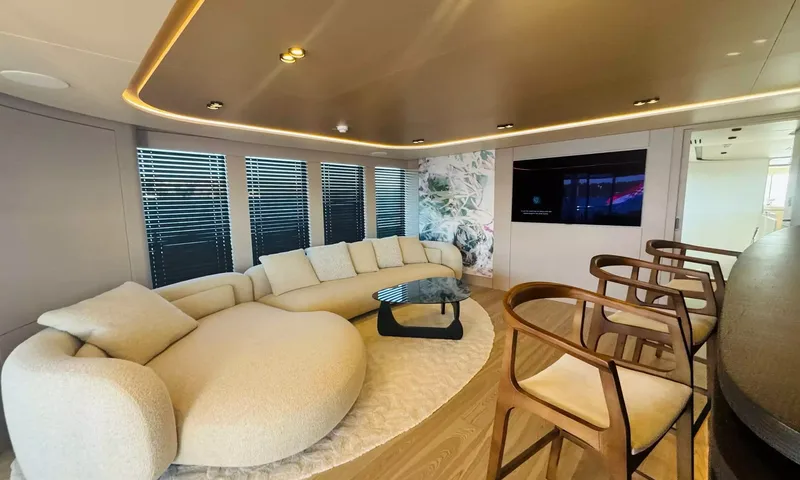 Oriy: Lounge com sofá curvo cor de creme, mesa de centro de vidro, televisão na parede e banquetas de bar de madeira