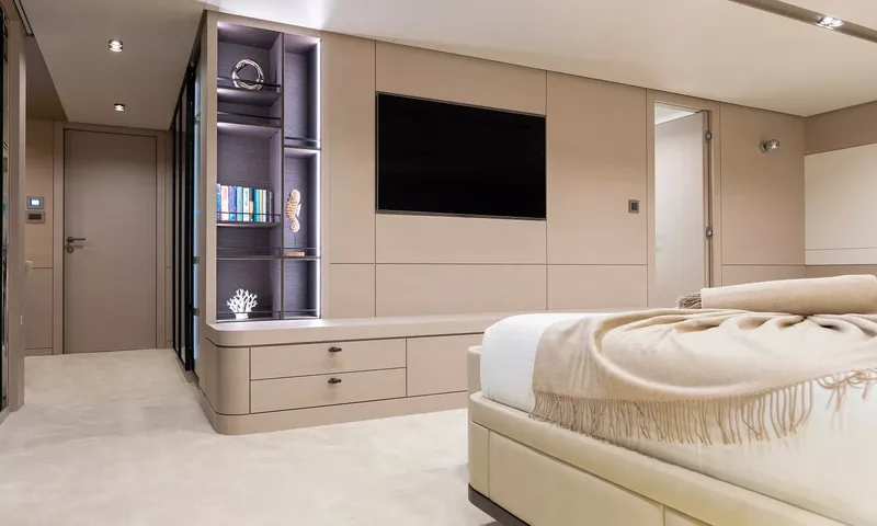 Oriy: Canto de cabine com prateleiras iluminadas, televisão na parede, gavetas e o pé de uma cama