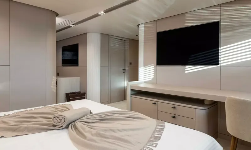 Oriy: Vista de uma cama em direção a uma escrivaninha, uma televisão na parede e uma porta, com luz do sol nas paredes