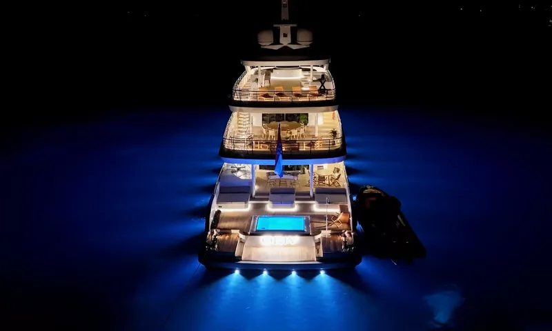 Oriy: Popa do iate à noite vista de cima, conveses iluminados, piscina na popa e luzes subaquáticas azuis