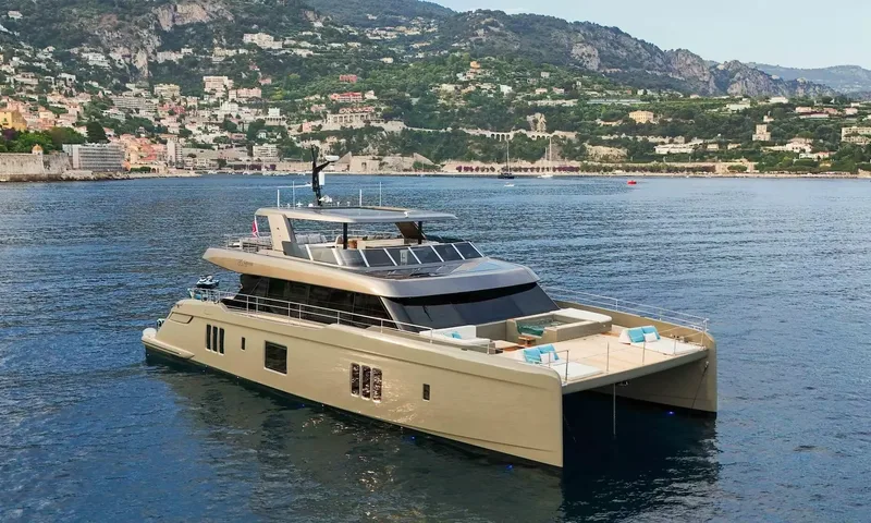Amitatu: Champagne colored power catamaran at anchor in a bay with a hillside town behind
