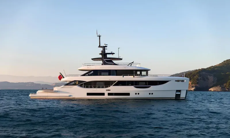 Aitana: White motor yacht at anchor in calm blue water near a green rocky coastline at dusk
