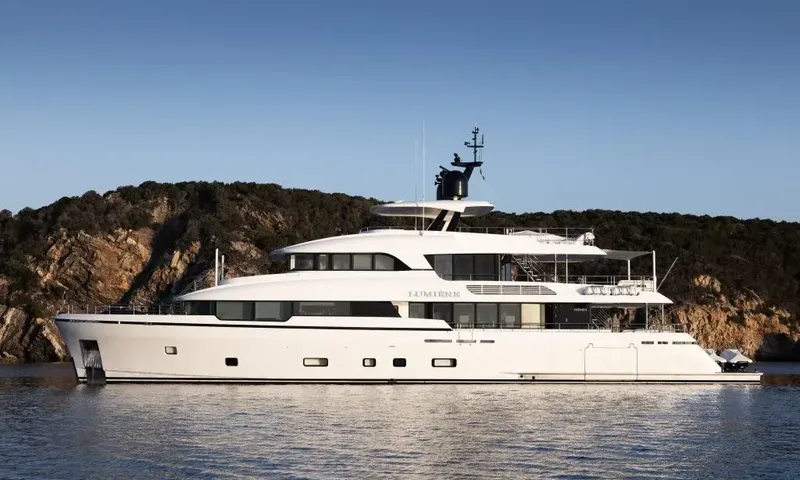 Lumière: Profile view of a white yacht anchored in front of a wooded rocky hillside