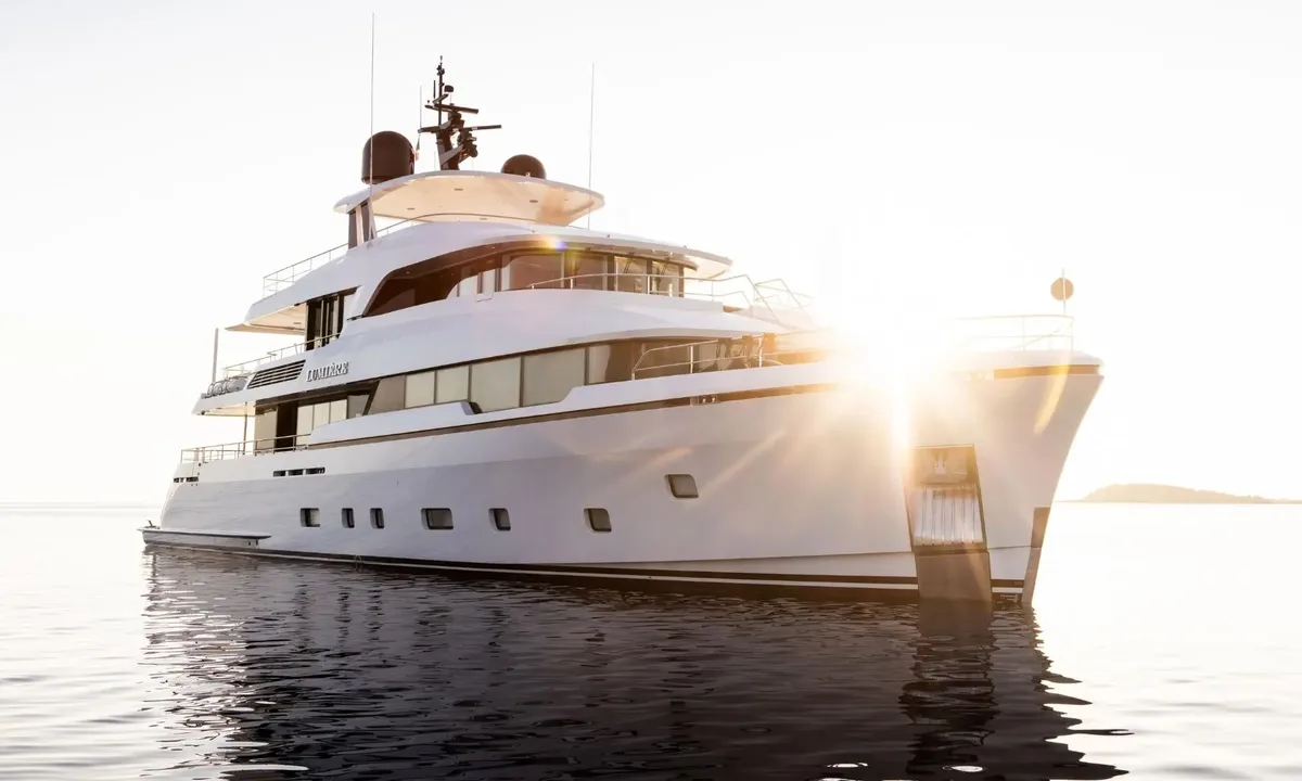 Lumière: White yacht at anchor on calm water with the low sun flaring behind its bow
