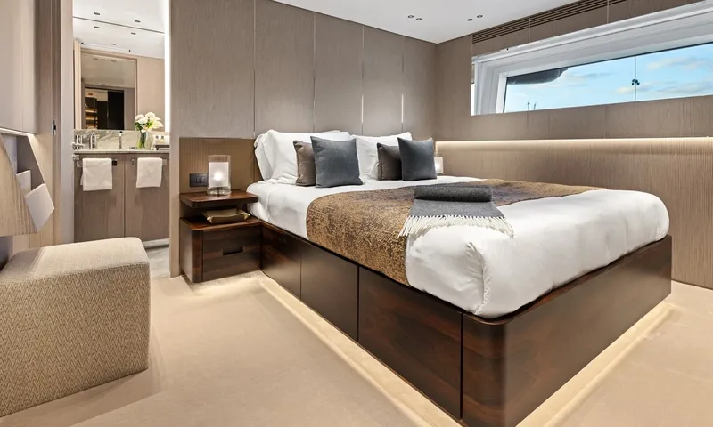 Wavė: Gästekabine mit Doppelbett, grauen Kissen, Holzbettsockel und Blick ins eigene Bad