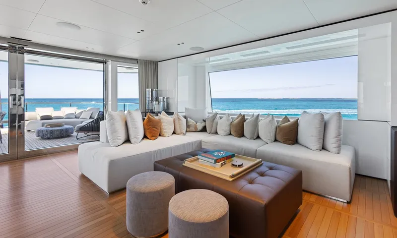 Wavė: Ecksofa mit Kissen, Lederpouf und Hockern vor einem breiten Fenster zum Meer
