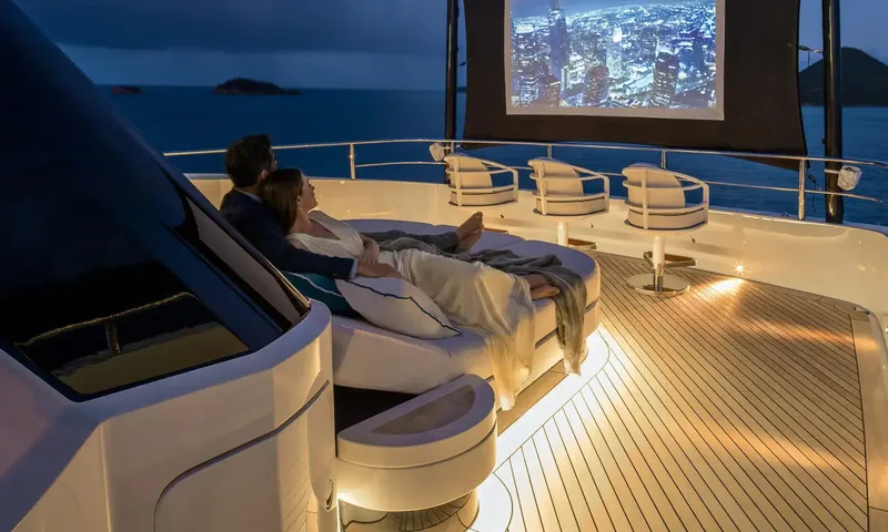 Amor à Vida: Couple reclining on a deck lounger at dusk watching a large outdoor screen beside bar stools