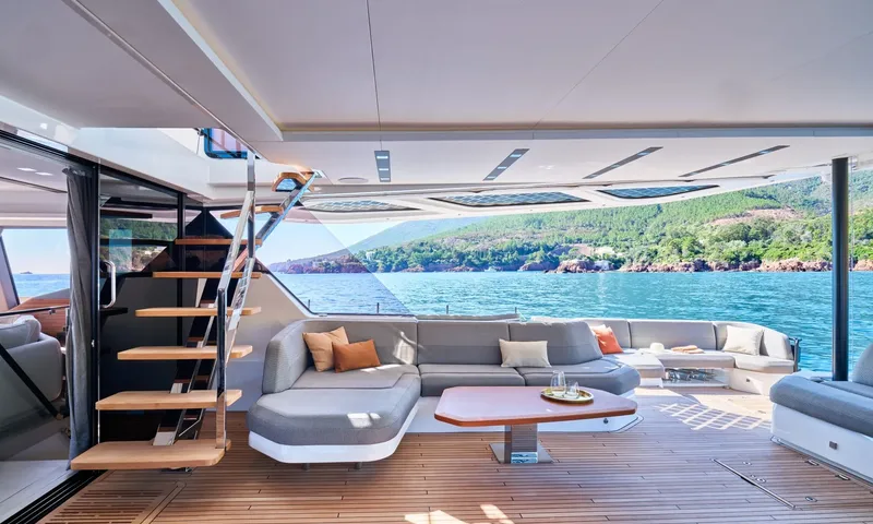 Alina: Shaded aft deck lounge with grey sofa, wooden table and stairs, overlooking a green coastline