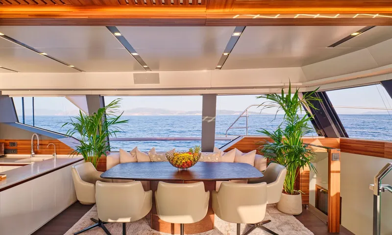 Alina: Indoor dining table with cream chairs, fruit bowl and palm plants before wide sea view windows