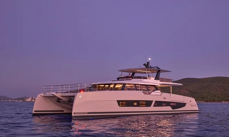 Alina: White power catamaran at anchor at dusk with lights glowing, hills behind