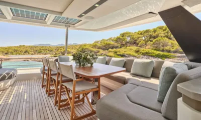Alina: Shaded outdoor dining table with wooden chairs, flowers and bench seating overlooking a green coast